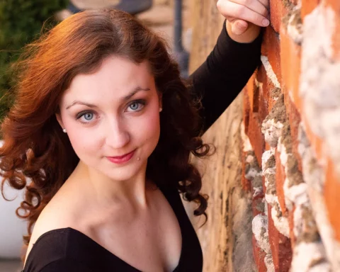 Close-up photo of an attractive young woman with red hair, green eyes, and fair skin leaning against a brick wall.