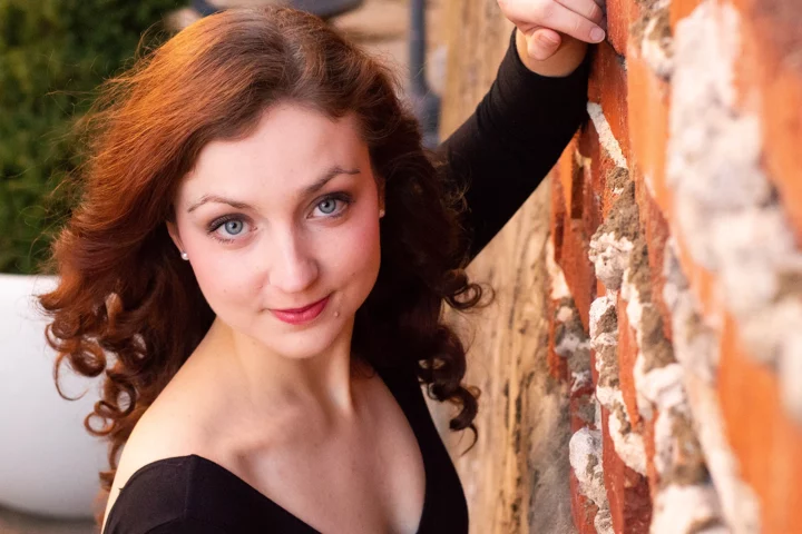 Close-up photo of an attractive young woman with red hair, green eyes, and fair skin leaning against a brick wall.