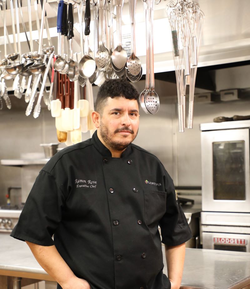 A young man, Chef Ramon Rosa, wearing a black chef's coat, standing in a commercial kitchen surrounded by  stainless steel appliances, counters, and utensils.
