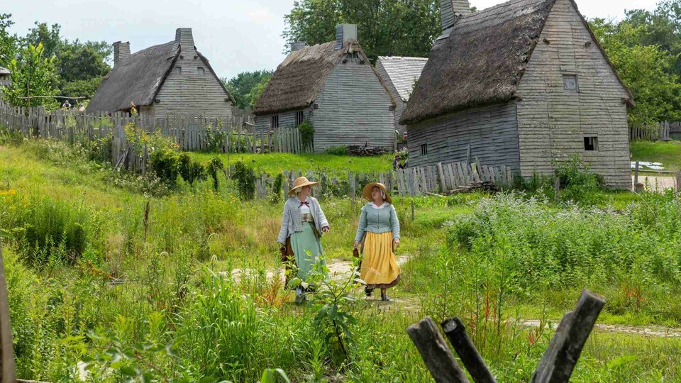 Two women in dresses stand among the green fields of Plimoth Patuxet Museum, with classic thatch houses in the background.